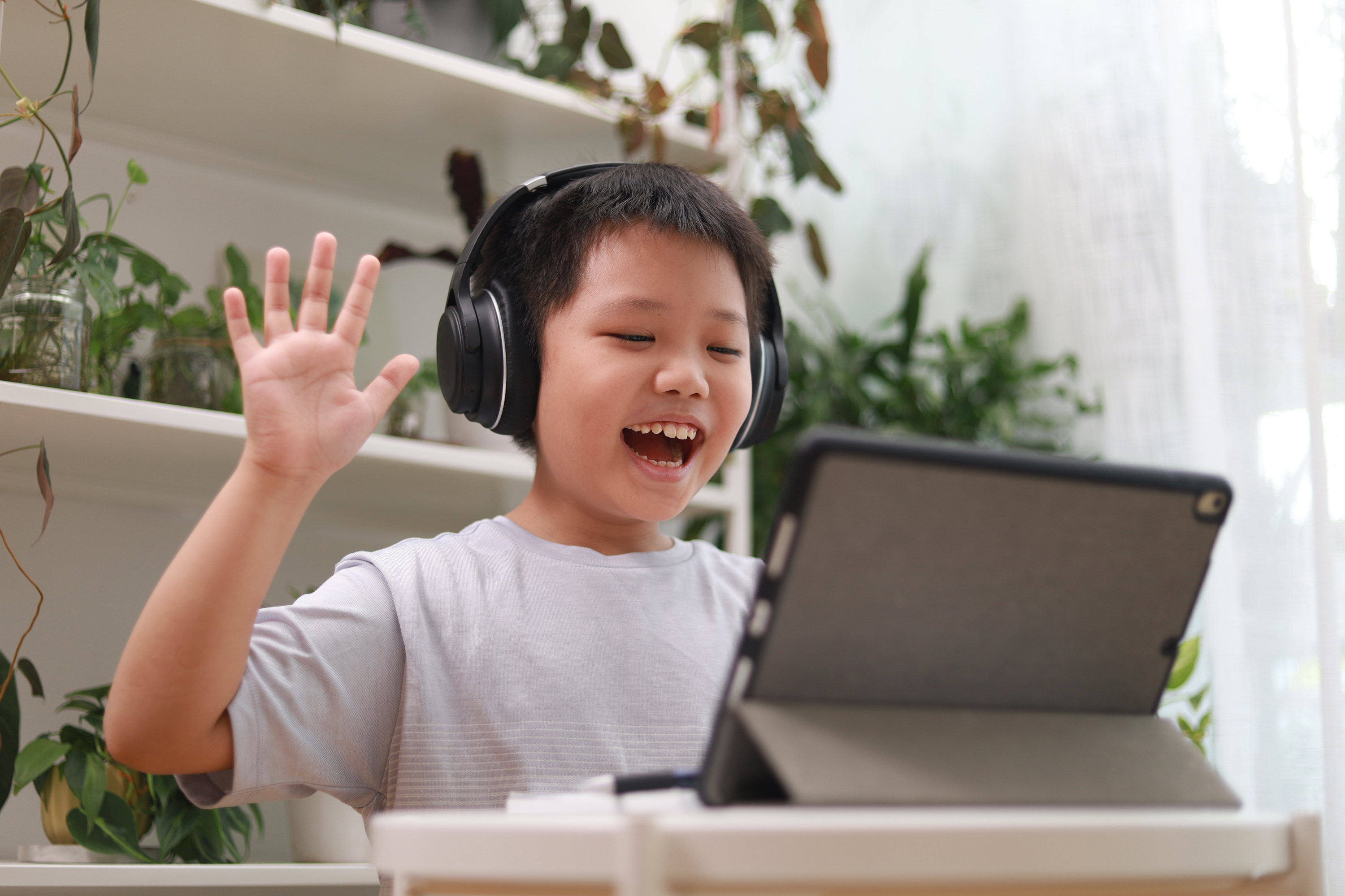 Young boy wearing headphones, smiling and waving at a tablet screen, with plants in the background.






